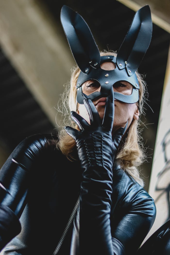 A woman wearing a black rabbit mask and a latex costume poses dramatically with hand on face.