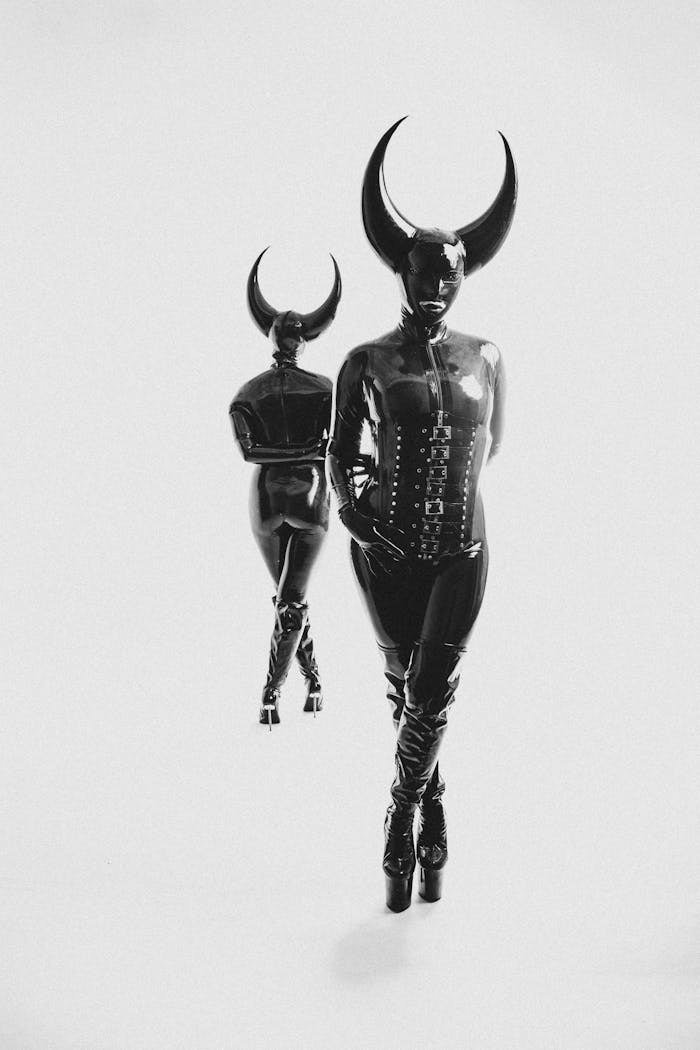 team-04 Two individuals in black latex costumes with horns and heels on a white background.